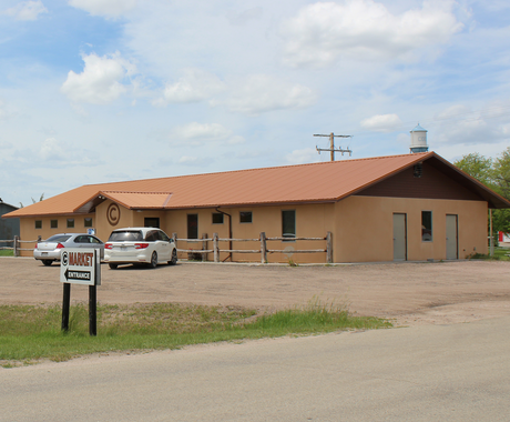 A tan, one-story commercial building with a reddish-brown metal roof and wooden fence sits beside a gravel parking lot. Two white vehicles are parked in front of the building, which has a round "C" logo above the entrance. A sign in the foreground reads "C Market Entrance." The sky is mostly clear with scattered clouds, and a water tower labeled "CODY" is visible in the background.