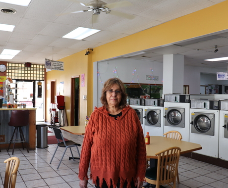 Mujer latina con gafas y una sudadera de manga larga color naranja se para en el centro de un negocio de lavandería con lavadoras y mesas con sillas por detras