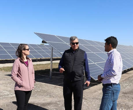 Three people standing in a field. Behind them are lines of solar panels and the people are conversing in conservation
