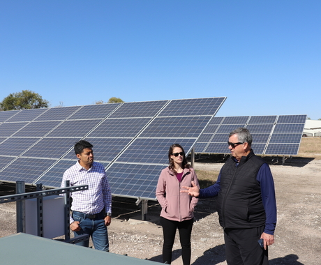 Three people standing in front of solar panels in the middle of a community solar array. 