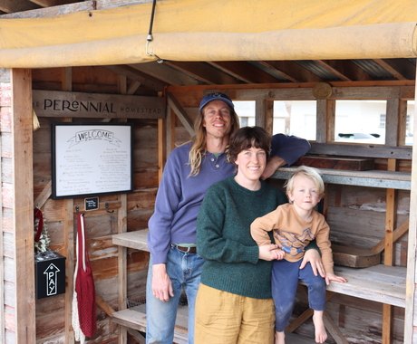 A man and woman stand in front of empty shelves in a wooden lean-to. The woman is holding a small boy sitting on one of the shelves. A sign that says "Perennial Homestead" is behind them.