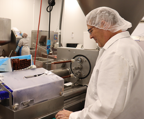 A man wearing a white coat and a white hair net stands in front of a stainless steel machine in a meat packing plant.