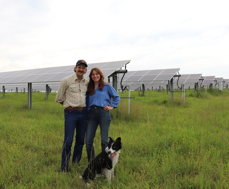 Man and woman standing in a field of longer grass. A black and white dog is in front of them. Solar panels are behind them. 