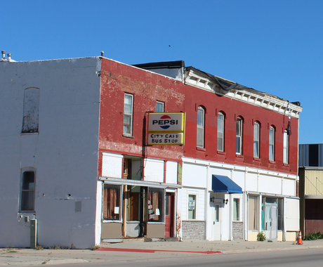 An old, two-story brick building sits along a highway. It appears to be vacant. On the upper half, we see red brick and a line of windows. The ground level has painted white brick. There is an old sign that says "PEPSI CITY CAFE BUS STOP."