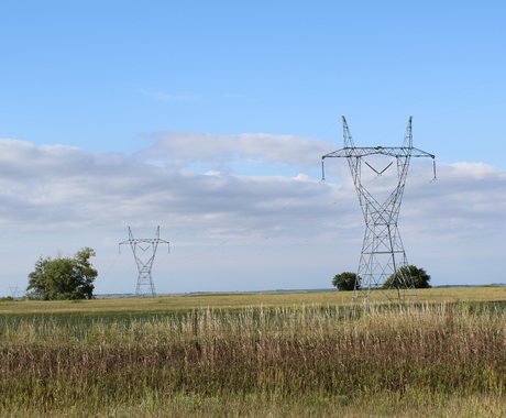 High-voltage power lines and transmission towers stretch across a flat, grassy rural landscape under a blue sky with scattered clouds.