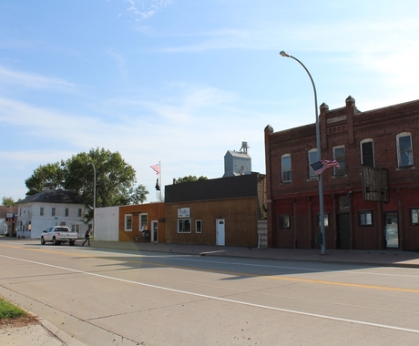 A row of downtown brick buildings in a small town, with the building to the far right a 2-story auditorium style. A gray grain elevator rises up behind the buildings.