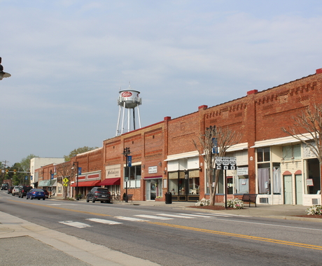Photo of downtown Halifax, VA, population 1,079. Showing one side of a block, with a highway running in front of it. The buildings are of red brick and white plus glass storefronts. A gray water tower is in the background with a red oval with "Halifax" in white lettering.