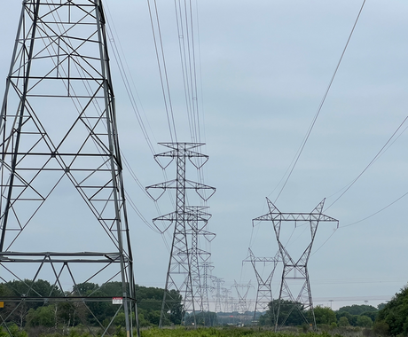 Tall metal transmission towers with power lines stretching into the distance across a green field under a cloudy sky.