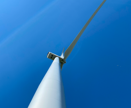 Close up shot of a white wind turbine, looking up toward the motor and propellers. The sky is bright blue in the background. 