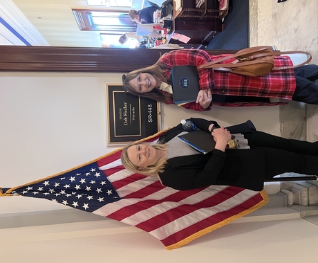 Dos mujeres de pie frente a un letrero, fuera de una oficina, que dice «Deb Fischer Nebraska», junto a una bandera estadounidense.
