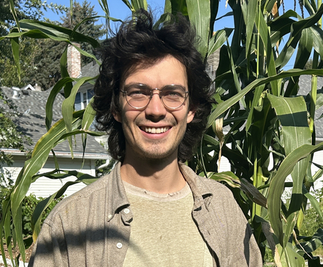 Man with shoulder length black hair and glasses wearing light green pants and shirt, with a long sleeve tan shirt over, stands in front of some corn 