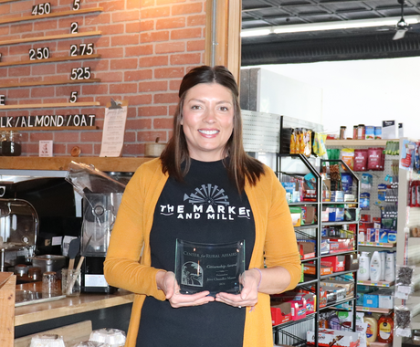A woman standing inside a store, smiling and holding a glass award. She is wearing a black 'The Market and Mill' T-shirt and a mustard yellow cardigan. Behind her is a coffee counter with a menu board, coffee machine, and baked goods on display. Shelves stocked with various grocery items are visible in the background.