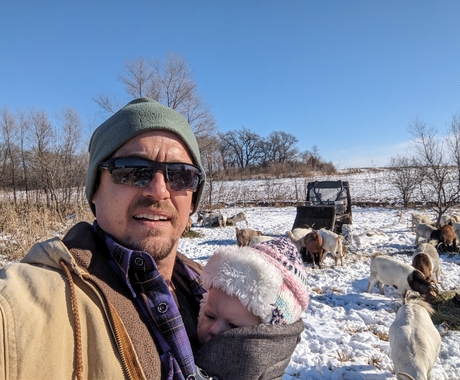 Man in a brownish coat and green stocking cap holding a baby wrapped in a dark blanket with a pink and white hat. Behind him is a snow covered field with goats grazing on hay he laid out.