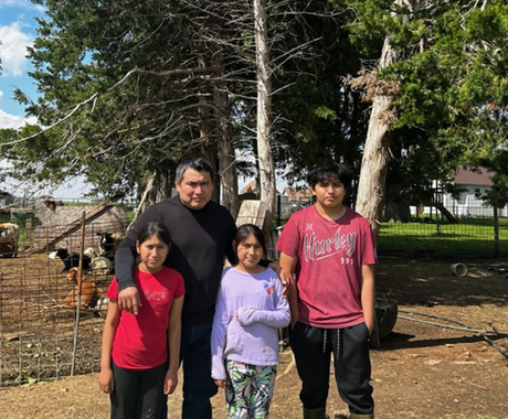Latino family with two girls, one boy, and a dad, poses in front of a pen with goats and pine trees 