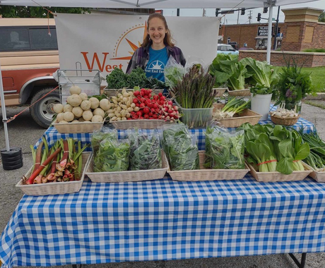 A woman with shoulder-length, light brown hair stands behind a folding table at what appears to be a farmers market. The table is covered with a blue and white checkered table cloth and holds wicker baskets of vegetables including asparagus, lettuce, green onions, and turnips.