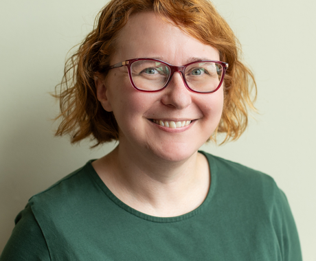 Woman with short red hair and red glasses smiles at the camera. She's wearing a forest green shirt and stands in front of a cream colored background.
