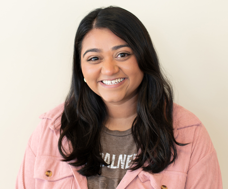 Woman with mocha colored skin and black long hair smiles at the camera. Woman is wearing a brown T-shirt with a pink jacket over the shirt.