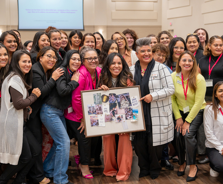 Picture of attendees at the 2024 Latina Women in Business Conference. They are all smiling, huddled around a young Latina women holding a vision board. She has long, dark hair, and smiles at the camera. 