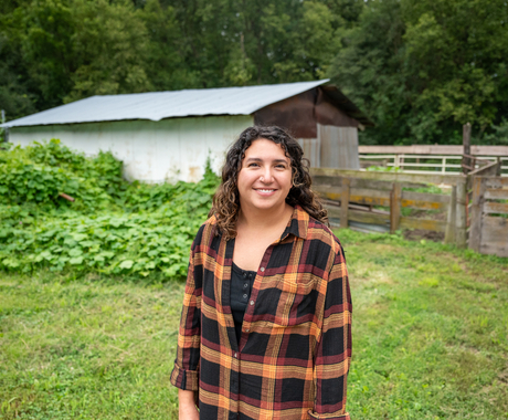 Woman with dark hair smiling at the camera, wearing an orange and black plaid long sleeve shirt. Background is an outbuilding with a wooden fence in front and a vine plant on the side.