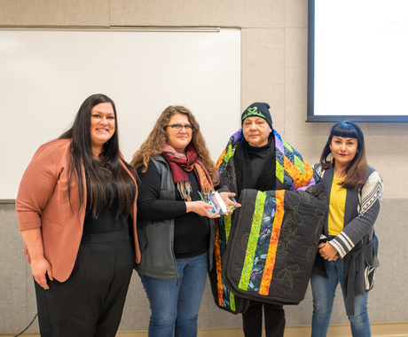 Four women smiling at the camera, two holding an award, one holding a Native star quilt