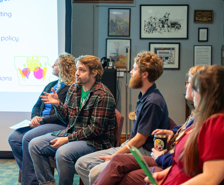 Five people sit in chairs with heads turned toward a screen. A man in a plaid button-down shirt appears to be addressing the room. 