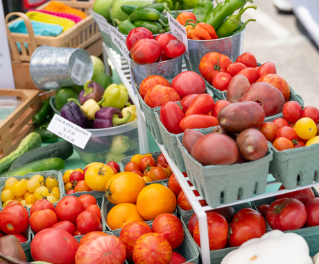 Colorful display of fresh produce at a farmers market, including a variety of heirloom tomatoes in shades of red, yellow, and orange; green bell peppers; chili peppers; and cucumbers. The produce is arranged in tin buckets and green cartons.