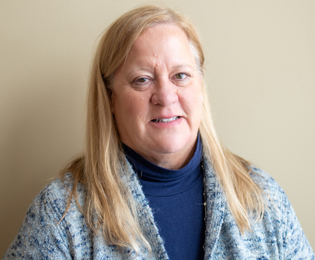 White woman with blonde hair wearing a blue turtle neck and a cardigan with blue and white standing in front of a beige wall