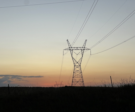 Fotografía de una torre de transmisión eléctrica al atardecer. La torre, los cables y el suelo que hay debajo se ven negros, con un cielo de color salmón a azul grisáceo detrás. No se aprecian otras estructuras en la foto.