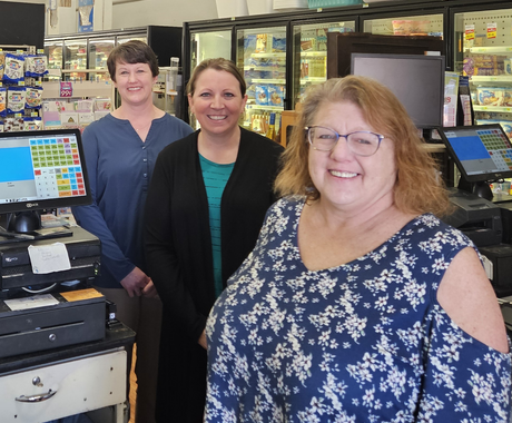 Three white women stand next to a grocery store cash register and smile at the camera. The woman in the back, to the left, has short brown hair and wears a blue long-sleeved shirt. The woman in the middle has brown hair pulled back into a pony tail and wears a teal top with a long black cardigan over it. The woman in the front and to the right has light brown, shoulder-length hair, blue glasses, and a blue floral top.