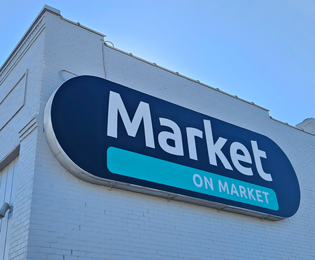The outside of the Market on Market building. It is a brick building painted cream/beige with a black oval sign that says "Market on Market."