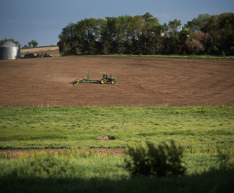 Un tractor tirant d'una jardinera, plantant en un camp nu de terra marró. Amb una zona d'herba en primer pla i una línia d'arbres a l'altre costat del camp.
