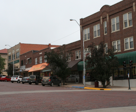 View of McCook, Nebraska Main Street buildings. On the far left, there is a large light-colored brick building, next to it is a dark brick two-story building, and then another two-story brick building with an orange awning. To the right, there are two more two-story dark brick buildings, each with dark brown awnings. To the right of those buildings is a three-story dark brick building with a dark green awning. Vehicles line the street in front of these buildings.
