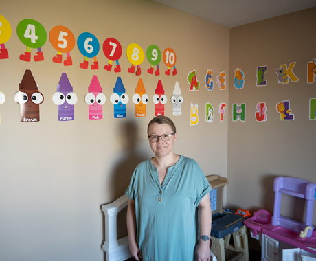 Woman in a short sleeved blue shirt, with pulled back dark hair, and glasses, smiles at the camera. She is standing in front of a beige wall with children toys stacked up behind her. On the wall behind her are colorful numbers, crayons, and letters.
