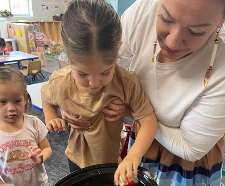 Una mujer sostiene a una niña para que añada ingredientes a una olla de cocción lenta. La olla tiene patatas.