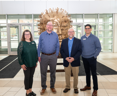 A group of four professionally dressed individuals stands smiling in a well-lit lobby in front of a large, sculptural golden head with raised features and metallic elements. The background features glass doors and large windows, allowing natural light to fill the space.
