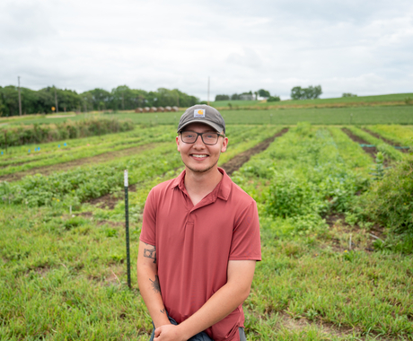 A young man stands in front of a green field. He is wearing a gray hat, glasses and a red polo shirt, with his hands folded in front of him.