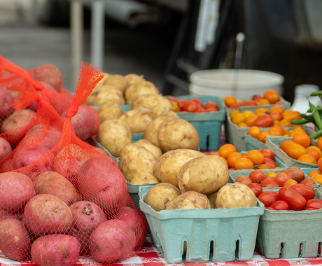 Productos frescos exhibidos en un mercado de agricultores, incluidas papas rojas en bolsas de malla, papas rojizas, tomates cherry y judías verdes dispuestas en cajas de cartón sobre un mantel a cuadros rojos y blancos.