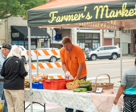 People shop at an outdoor farmer’s market under a canopy tent. A vendor in an orange shirt arranges produce on a table, which displays fresh vegetables like zucchini and cucumbers. Several shoppers stand in line, and buildings and cars are visible in the background.
