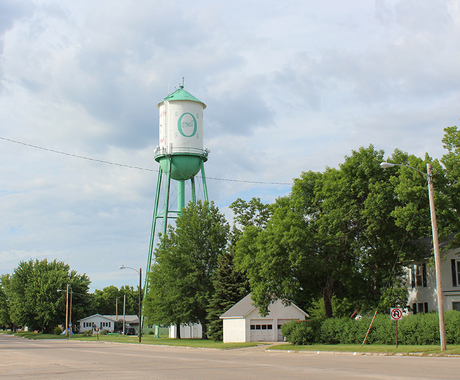 Water tower that's white with a green top and green bottom, on a street with mature trees and houses