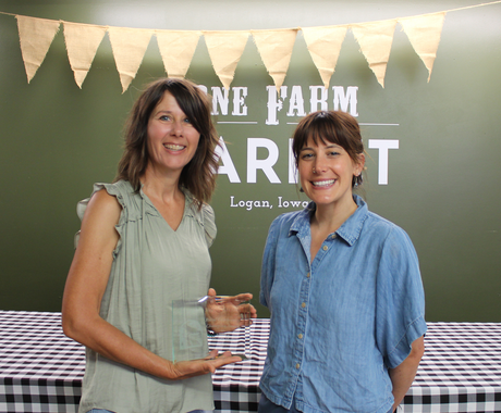 Two women stand side by side, smiling at the camera. The woman on the left has shoulder-length brunette hair, a sage-colored sleeveless top, and holds a rectangular glass award. The woman on the right has brunette hair with bangs, swept back in a ponytail. She wears a short-sleeve chambray button-down shirt. Behind them, there is a table with a black and white plaid table cloth and a green wall that says "One Farm Market Logan, Iowa" with burlap pennants hanging from the ceiling.