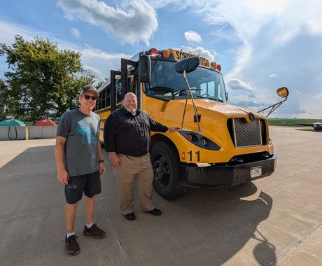 Two men stand to the front and passenger side of a yellow school bus with a blue sky and fluffy clouds above.