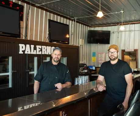 Two men standing on either side of a shiny metal bar, with "Palermo" behind the bar and three TVs on the wall that are turned off. The two men are wearing black shirts and ball caps.
