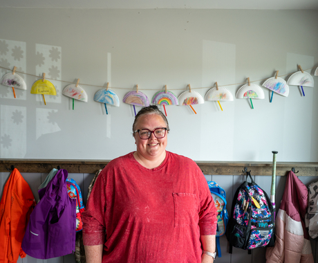 Woman with a 3/4 sleeve dark pink shirt with a pocket, looks at the camera. Her hair is pulled back and she's wearing glasses. She stands in front of a wall of hooks for little one's coats and backpacks. Above the hooks is a string of half paper plate bright constructions.