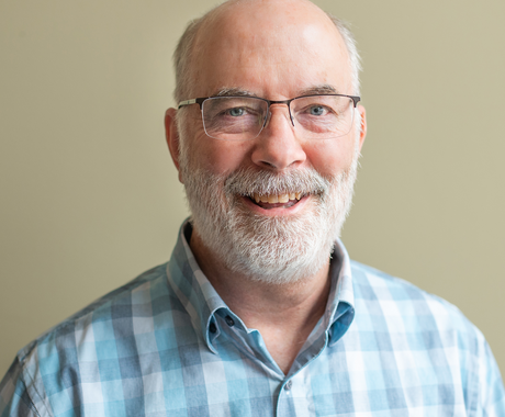 Smiling older man with a bald head, white beard, and glasses, wearing a light blue plaid shirt, standing in front of a neutral-colored background.