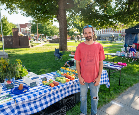 Man dressed in red T-shirt and jeans with a hole at the knee. Man is standing in front of a table covered in a blue checkered tablecloth, full of veggies including dill, tomatoes (in red, orange, green), and more veggies. In the background is a grassy area, then downtown buildings.