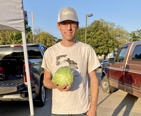 Man in ballcap, T-shirt, and jeans holding a lettuce, looking at the camera. In the background are the backs of 2 pickup trucks and the leg of a pop-up tent.