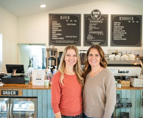 Two women in front of a counter where you order coffee, with a menu board behind them