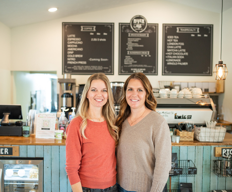 Rachel Barlow and Heather Veik stand in front their coffee bar with three menu boards in background