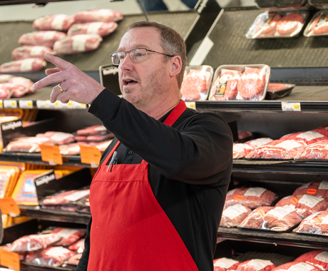 A man stands in front of a grocery store meat cooler with his left hand raised, appearing to be speaking. He wears glasses, a black long sleeve shirt, and a red apron. 