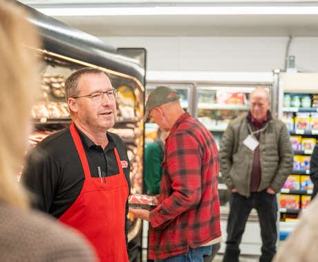 Man with short, brown hair and glasses stands in front of a grocery cooler wearing a black polo shirt and a red apron over it.